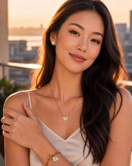 A woman with long dark hair wearing a gold heart pendant necklace, matching bracelet, and stud earring, posing outdoors during sunset with a blurred city skyline in the background.