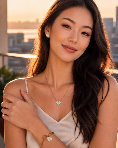 A gold heart-themed jewelry set displayed on a white pedestal, featuring a gold chain necklace with a split gold and light blue heart pendant, a gold link bracelet with matching heart charms, and small heart earrings. A white frosted cake with colorful sprinkles sits in the softly blurred background against a muted blue-gray wall.