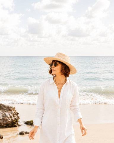 A woman wearing a wide-brimmed straw hat, sunglasses, and a long white button-up shirt walks along a sandy beach with the ocean and cloudy sky in the background.