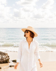 A woman wearing a wide-brimmed straw hat, sunglasses, and a long white button-up shirt walks along a sandy beach with the ocean and cloudy sky in the background.