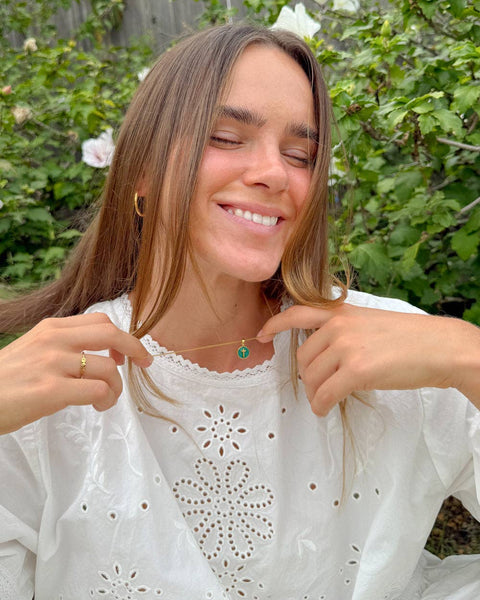 Woman wearing a white blouse with lace details, pointing to a necklace with a green pendant against a natural background.