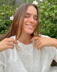 Woman wearing a white blouse with lace details, pointing to a necklace with a green pendant against a natural background.
