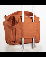An orange-brown woven fabric luggage bag with dual top handles and a wide wrap-around strap, shown attached to the telescoping handle of a white suitcase against a plain white background.