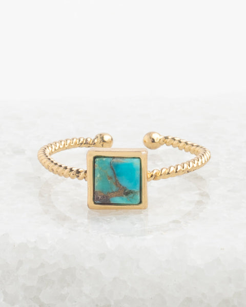 A gold-toned open bangle ring featuring a square turquoise stone inlay with natural matrix patterns, displayed on a bed of white salt crystals.