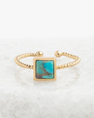 A gold-toned open bangle ring featuring a square turquoise stone inlay with natural matrix patterns, displayed on a bed of white salt crystals.