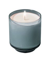 A lit scented candle in a ribbed, smoky gray glass jar with a white wax surface and a single flame, isolated on a white background.