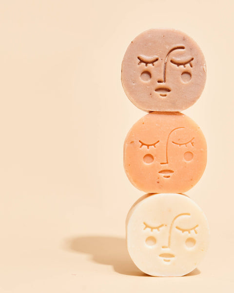 Three round, face-relief bath or body soaps stacked vertically on a warm beige background. The top soap is a muted dusty rose, the middle is a soft peach, and the bottom is an off-white cream.