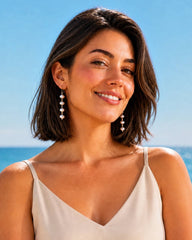 A woman with a short brown bob hairstyle wearing dangling pearl-like drop earrings and a cream camisole top, photographed outdoors against a bright blue sky and ocean background.