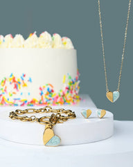 A gold heart-themed jewelry set displayed on a white pedestal, featuring a gold chain necklace with a split gold and light blue heart pendant, a gold link bracelet with matching heart charms, and small heart earrings. A white frosted cake with colorful sprinkles sits in the softly blurred background against a muted blue-gray wall.