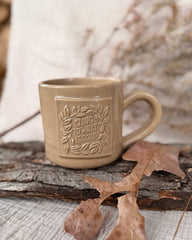 A beige ceramic mug with a handle and embossed floral border design featuring  CLING TO WHAT IS GOOD on the front, styled on a rustic log with dried leaves and a soft, blurred background.