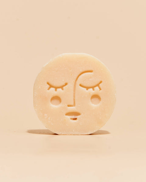 A single round, pale beige bar of soap with a simple embossed face design, standing upright on a plain beige background.