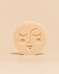 A single round, pale beige bar of soap with a simple embossed face design, standing upright on a plain beige background.