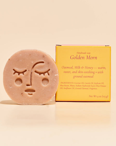 A handmade soap bar with a minimalist face design sits next to its yellow packaging box on a warm beige background. The box is labeled 'Golden Morn' and reads 'Handmade soap'.
