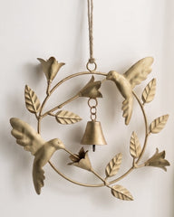 A brass bird wreath wall hanging with three birds, leaves, and a small bell, suspended by a rope against a white wall.