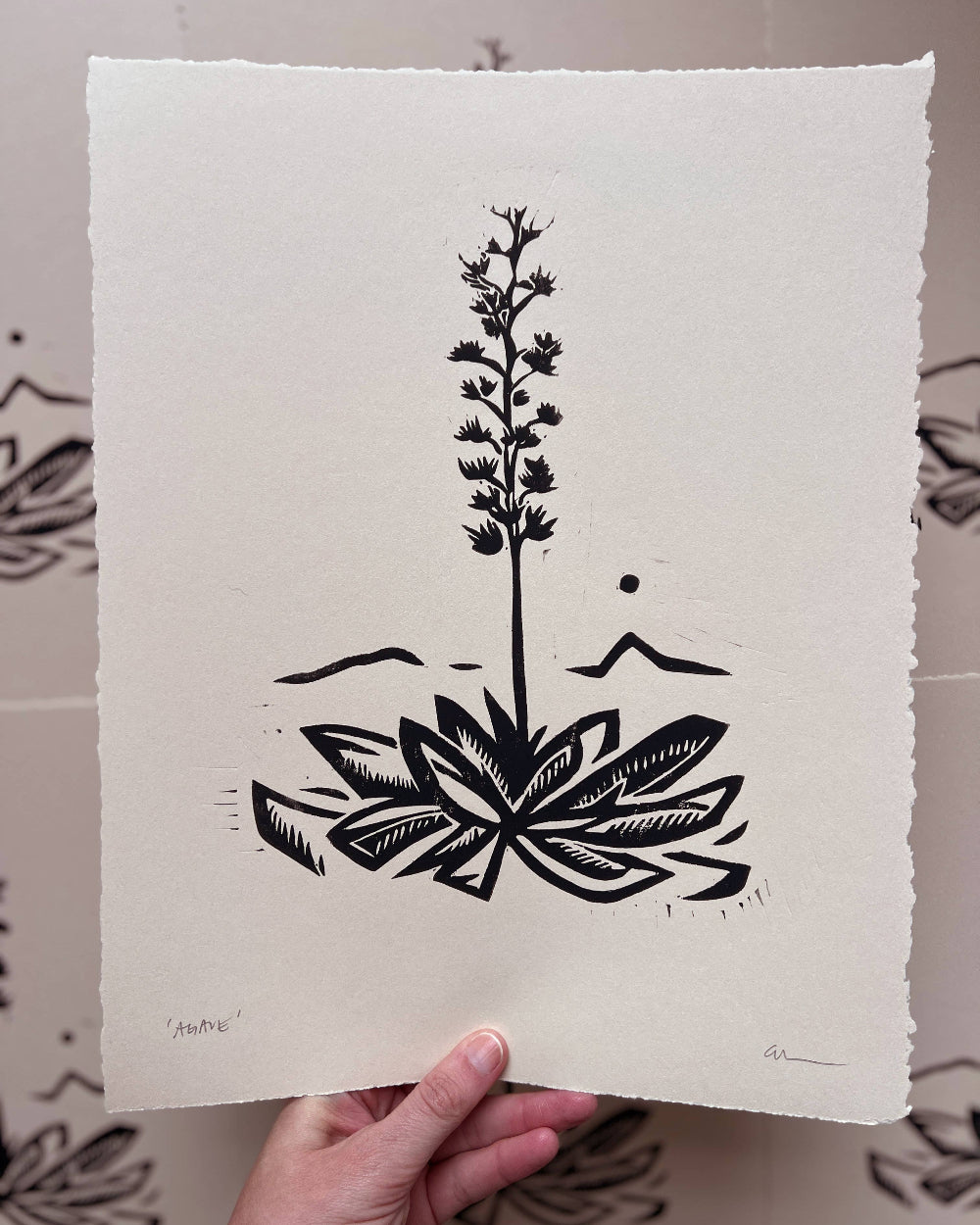 Hand holding a piece of paper with black line art of a plant design against a white background.