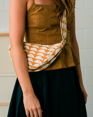 A woman wearing a mustard brown camisole and a dark skirt carries a patterned fabric crossbody bag with an orange and cream scallop design.
