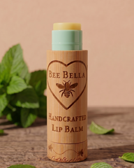 Bee Bella lip balm with wooden container on a wooden surface with green leaves in the background