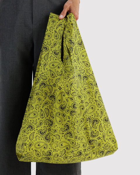 A person holding a large, soft-sided tote bag with a black paisley pattern on a bright yellow-green background, paired with dark gray trousers against a light gray studio backdrop.