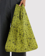 A person holding a large, soft-sided tote bag with a black paisley pattern on a bright yellow-green background, paired with dark gray trousers against a light gray studio backdrop.