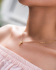 Gold necklace with a cross pendant worn by a person in a light pink top.