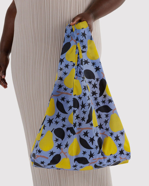 A person wearing a beige pleated dress holds a large fabric tote bag with a light blue background and a yellow pear, black leaf, and black star pattern.