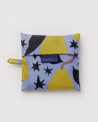 A small BAGGU reusable bag in a light blue fabric with a yellow, black, and brown abstract pattern, featuring a black flap closure and a blue brand label.