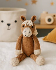 A knitted plush toy horse sitting on a white fabric surface in a softly styled room. The toy is brown with a cream-colored face, mane, and hooves.
