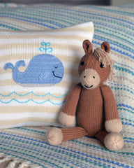 Knitted horse toy next to a pillow with a whale design on a striped blanket