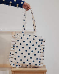 Tote bag with blue polka dots held by a person on a wooden stool against a white background
