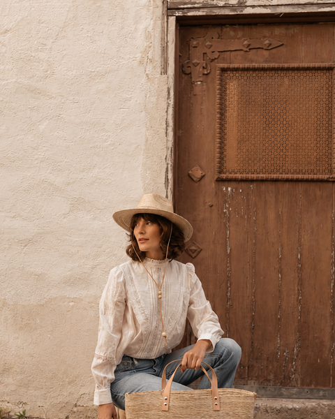 Woman in a white blouse and hat sitting against a textured wall with a wooden door.