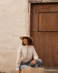 Woman in a white blouse and hat sitting against a textured wall with a wooden door.