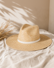 Beige straw hat with a white band on a beige surface with dried plants.