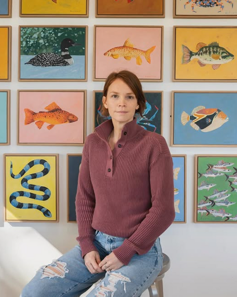 A woman wearing a ribbed maroon mock-neck sweater and distressed blue jeans sits on a stool in front of a gallery wall of colorful framed fish paintings.