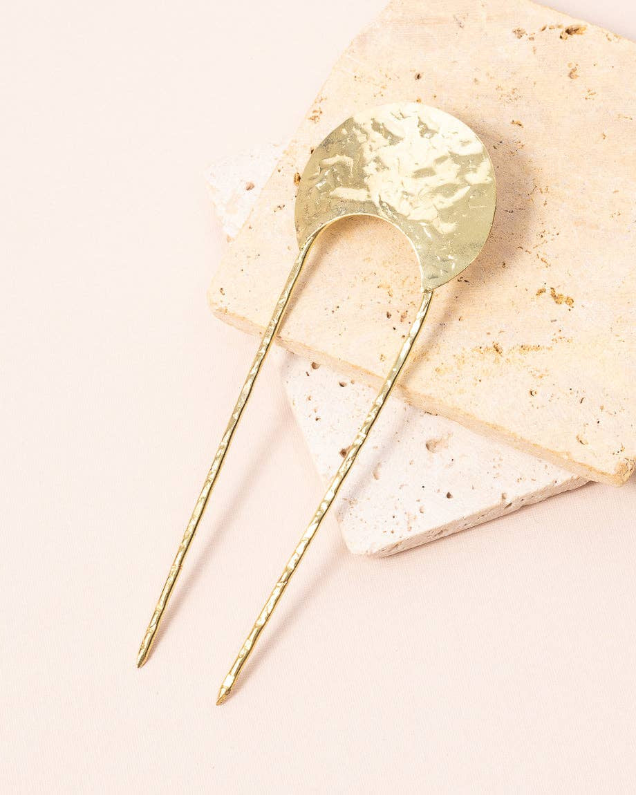 A hammered gold hair pin with two twisted wire prongs, displayed on a stack of textured beige and white stone tiles against a solid pale pink background.