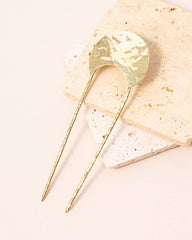 A hammered gold hair pin with two twisted wire prongs, displayed on a stack of textured beige and white stone tiles against a solid pale pink background.