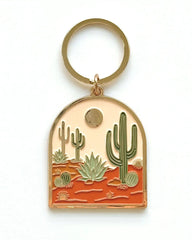 A gold-tone metal keychain featuring an arched pendant with an enamel desert landscape design. The design includes green saguaro cacti, a green agave plant, and a reddish-orange desert ground under a pale peach sky with a gold sun.