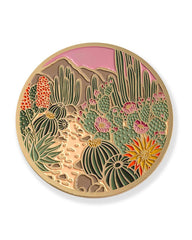 A round enamel coaster featuring a desert landscape with various green cacti and succulents, pink flowers, a sandy path, and a pink sky, all outlined in gold metal.