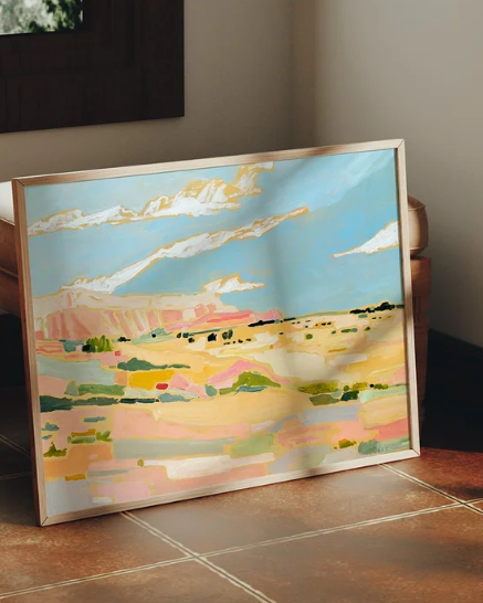 Framed painting of a landscape with a blue sky and pinkish ground on a tiled floor.