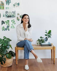 A woman sits on a wooden bench in a bright room, wearing a white button-up shirt, blue jeans, and white slip-on shoes. The background features a white wall decorated with botanical art prints, and several potted green plants are arranged around the bench.