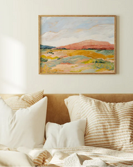 A framed landscape painting hangs on a cream wall above a bed with beige and white pillows and a textured cream blanket.