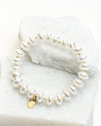 A pearl beaded bracelet with small gold spacer beads and a gold clasp, displayed on a white textured stone against a dark blue background.