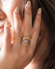 A close-up of a person's hand wearing three stacked rings: two textured gold rings and one silver ring with a small white stone, against a soft-focus background of skin and dark hair.