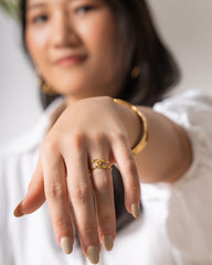 A close-up lifestyle shot of a woman wearing a gold ring on her finger, with her hand extended toward the camera. The background is softly blurred, showing a woman in a white blouse.