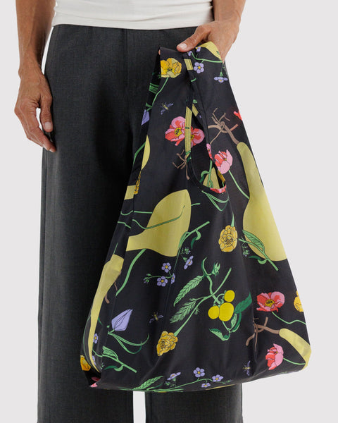 A person wearing dark gray trousers holds a large reusable tote bag with a black background and a colorful botanical print featuring yellow leaves, pink flowers, and green stems.