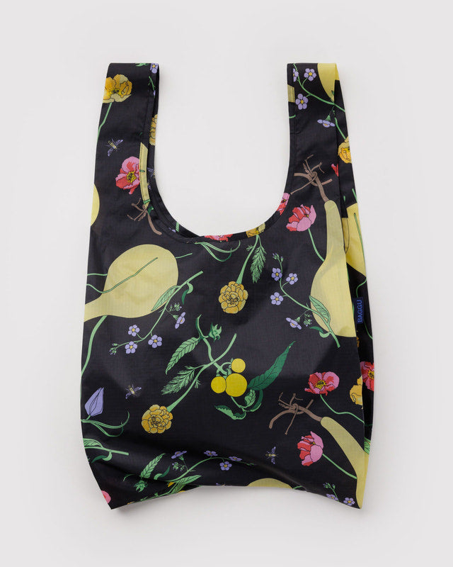 A reusable shopping bag with a black background and a colorful botanical print featuring yellow pears, green leaves, pink flowers, and small purple blossoms, shown against a plain light gray background.