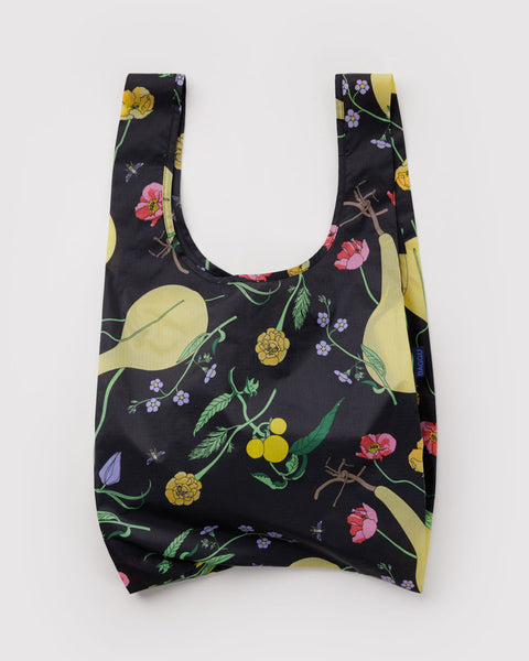 A reusable shopping bag with a black background and a colorful botanical print featuring yellow pears, green leaves, pink flowers, and small purple blossoms, shown against a plain light gray background.