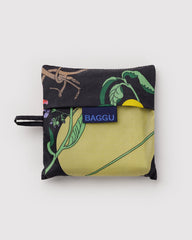 A small BAGGU reusable bag with a black background and a botanical print featuring green leaves, yellow accents, and a yellow-green leaf design. The brand name 'BAGGU' is printed on a blue fabric tag on the front.