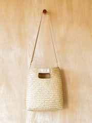 A natural woven straw shoulder bag with a long strap, hanging from a small wooden peg on a light wood wall.