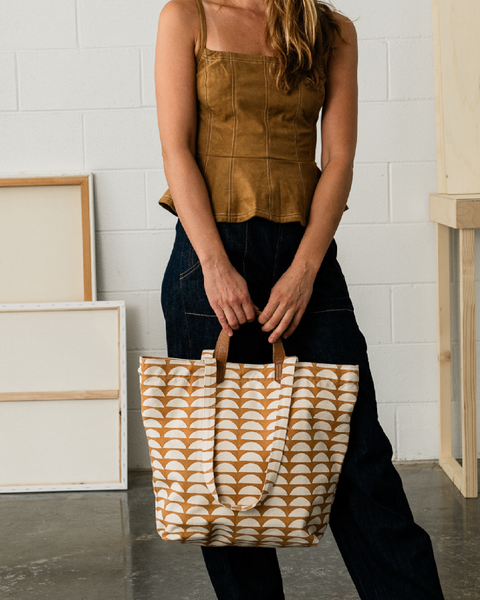 A woman wearing a brown sleeveless top and dark jeans holds a large cream canvas tote bag with a tan repeating wave pattern and brown handles in a studio setting.
