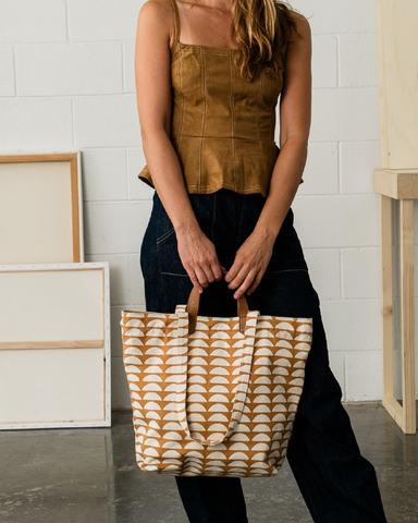 A canvas tote bag featuring a repeating ochre orange and cream semicircle pattern, with solid brown leather handles and trim, shown against a plain light gray background.